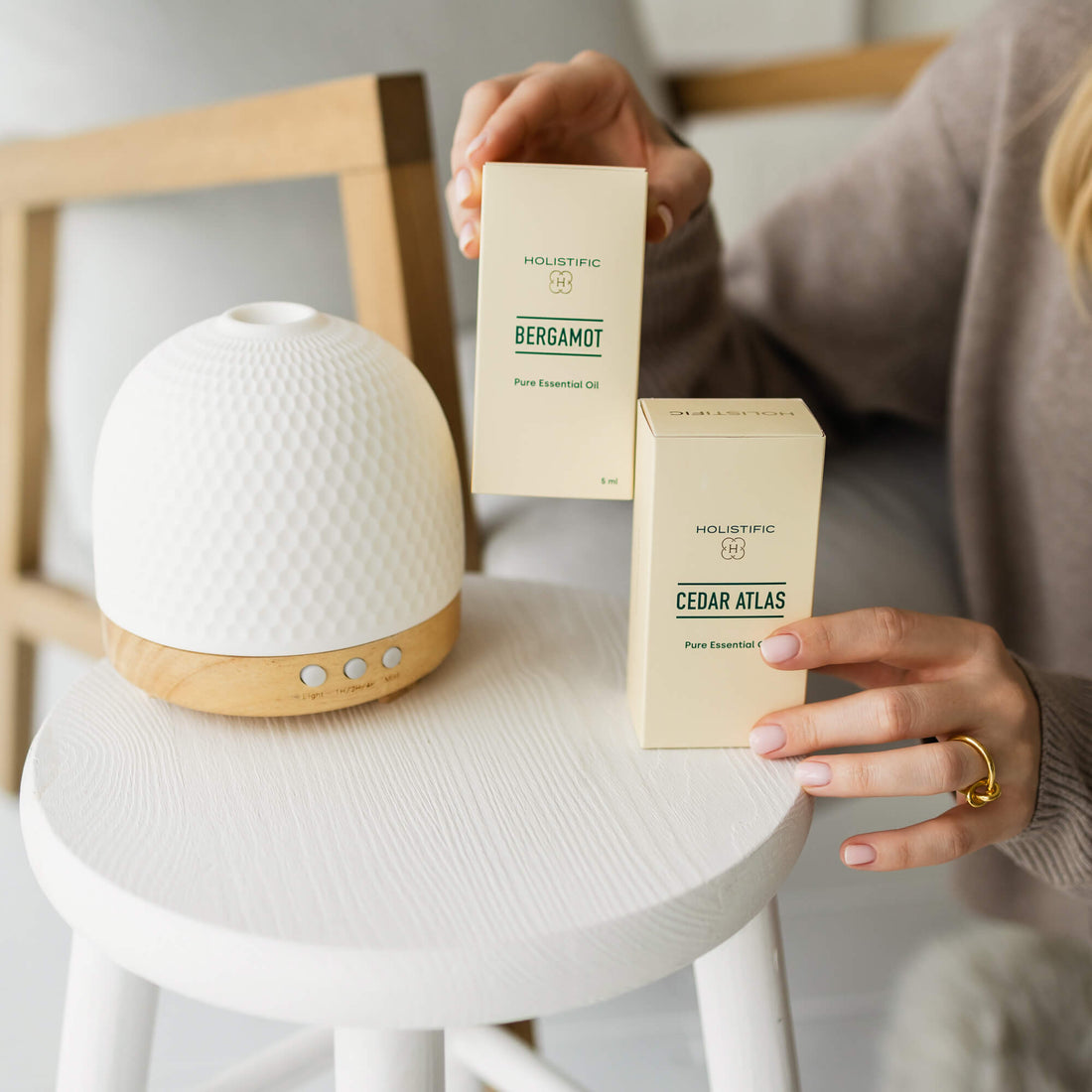 Difuzér Mellow v popředí. Ženské ruce drží krabičky s esenciálními oleji Bergamot a Cedr atlaský.